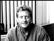 A black and white photo of a smiling man with tousled hair, seated, and wearing a casual sweater, about Sun Valley Construction, against a vertically striped background.