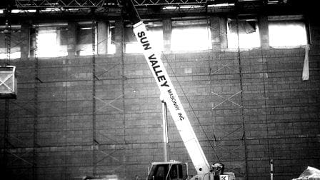 A black and white photo about Sun Valley Construction shows a construction site with a telescopic boom lift, labeled "Sun Valley," extending towards a partly constructed wall or building facade.