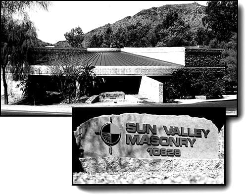 A black and white photograph displaying the entrance to a property with a sign that reads "Sun Valley Masonry 10629" in the foreground and a building with low-pitched roofs nestled among trees in