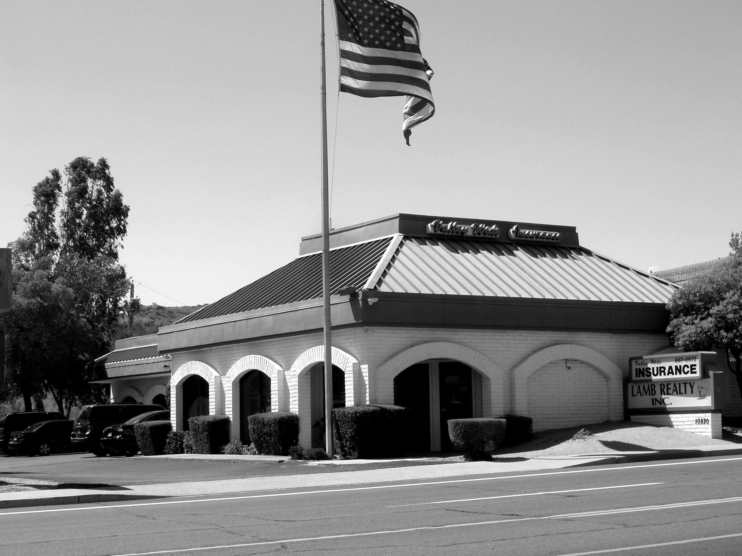 A monochrome photo of a single-story commercial building with an american flag flying overhead, and signs indicating the presence of an insurance and realty business, about Sun Valley Construction.