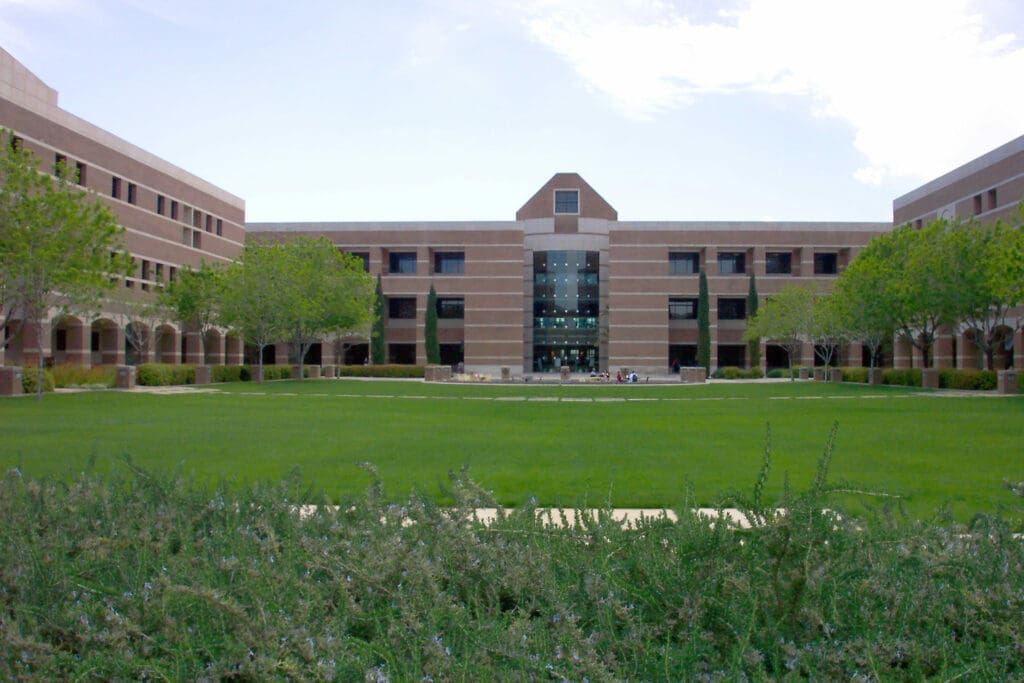 A symmetrical building with a central glass entryway, surrounded by well-maintained lawns and shrubs under a clear sky.