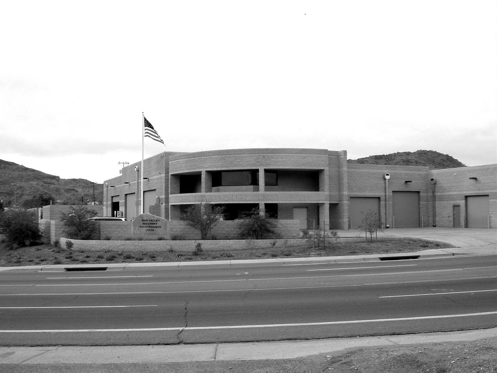 A grayscale image depicting a modern single-story building constructed by Sun Valley Construction, with a flagpole flying the American flag, situated next to a road with visible lane markings.