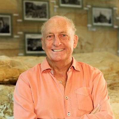 An elder man with a warm smile, wearing a salmon-colored shirt adorned with the logo of Sun Valley Construction, is sitting in front of a wall filled with framed black-and-white photographs.