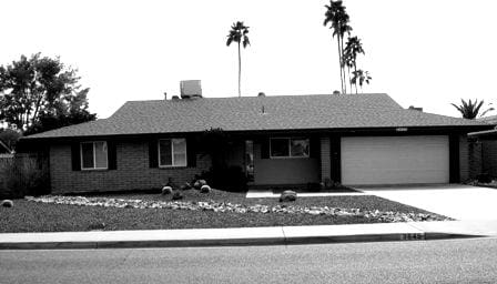 A black and white photograph of a single-story residential house, about Sun Valley Construction, with a shingle roof, a double garage door, and a landscaped front yard with palm trees.