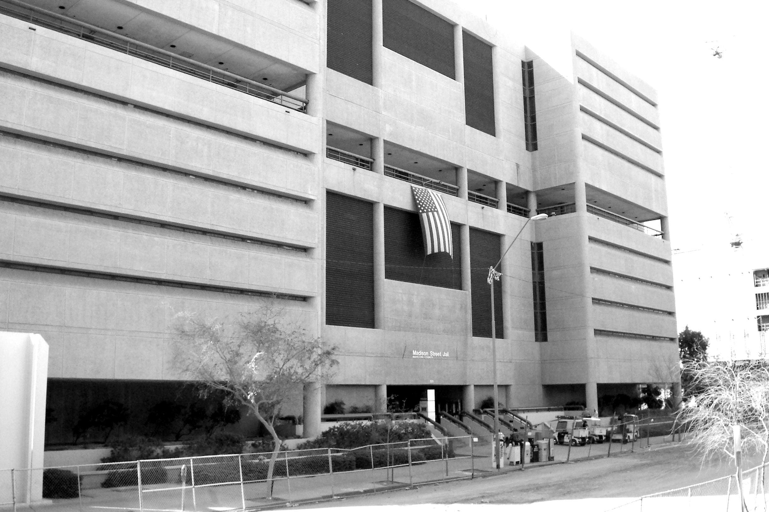 A black and white photo of a modern multi-story building about Sun Valley Construction with an American flag hanging on its facade, and a few trees and benches visible in front.