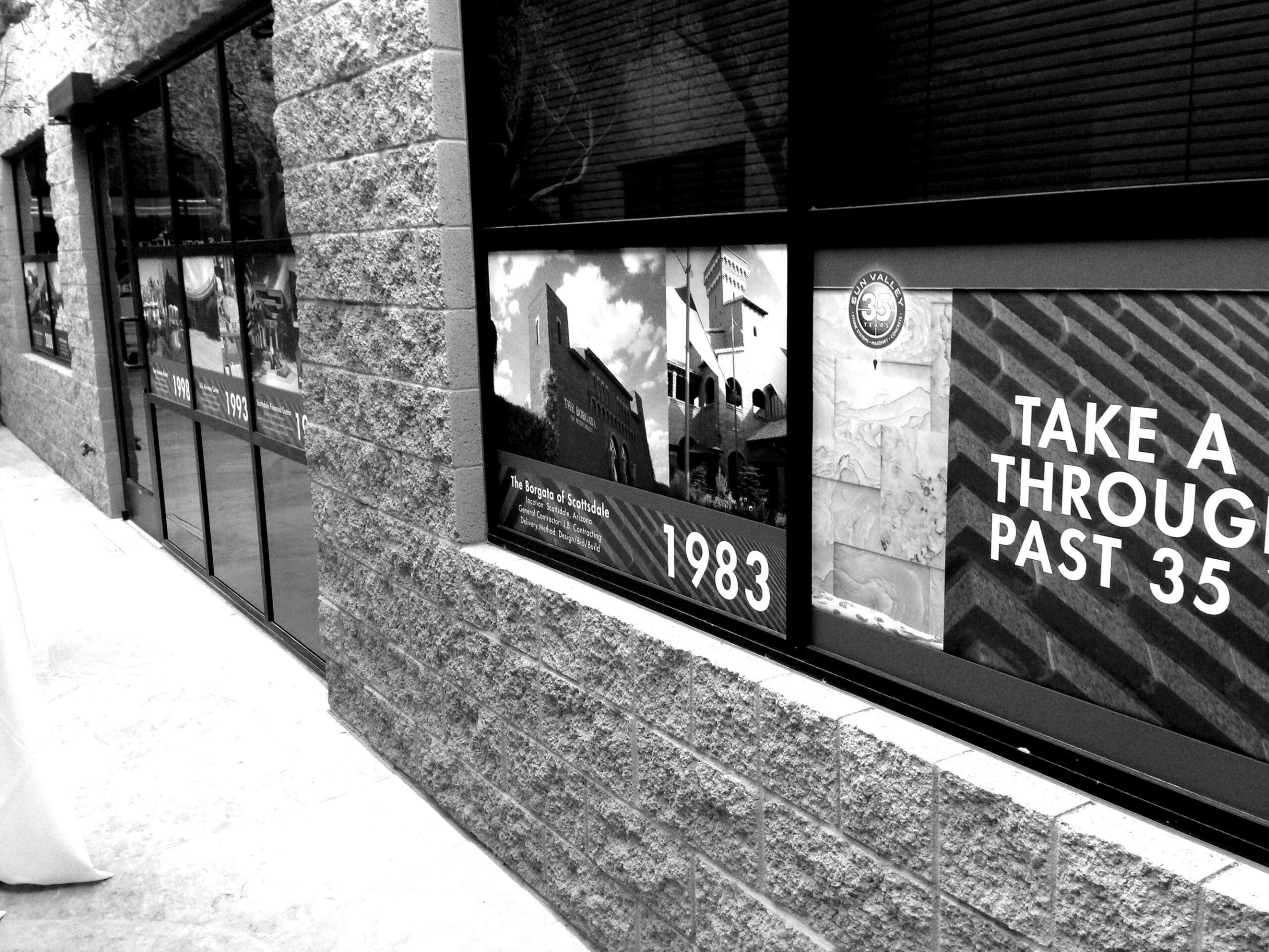 A black and white photo showing a building with large windows displaying an exhibition about Sun Valley Construction with texts and images; one text reads "1983", and another says "take a tour through past