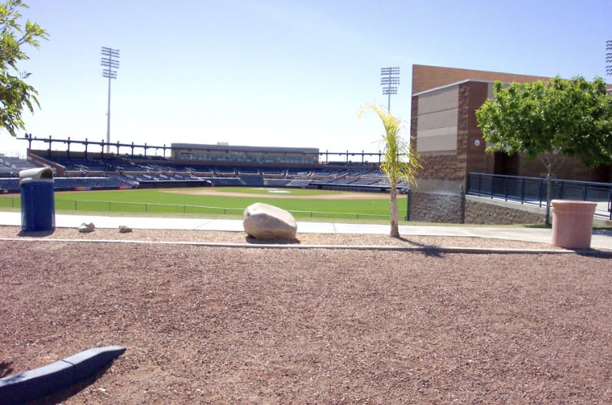 A daytime view of an empty baseball stadium seen from beyond the outfield, featuring a well-manicured field, stadium seating, and clear skies.