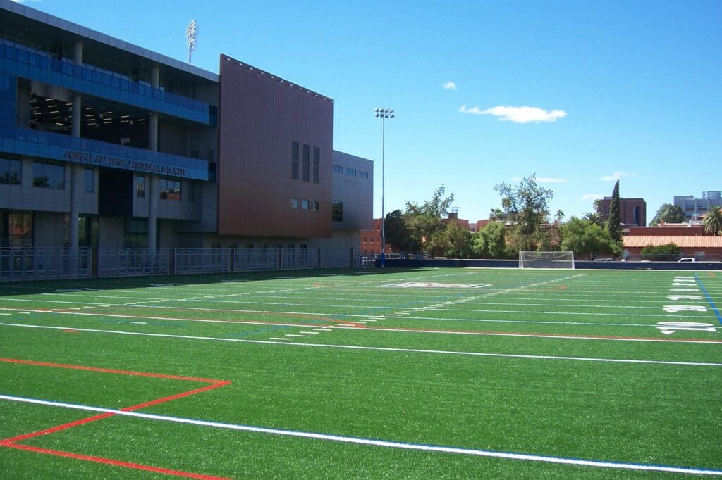 A clear day overlooking an empty artificial turf sports field with red and white boundary lines, adjacent to a multi-story building with signage.