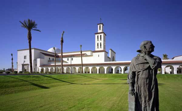 A statue of a person stands in the foreground, gazing toward a white building with a bell tower and multiple arches, surrounded by palm trees under a clear blue sky.