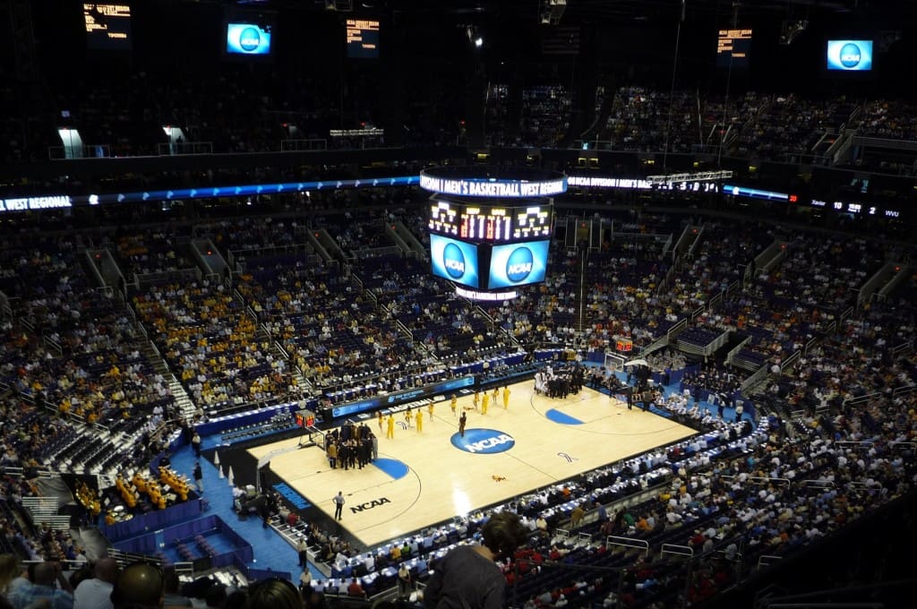 An overhead view of an indoor basketball arena during an ncaa tournament game, with teams gathered on the court and spectators filling the stadium seats.