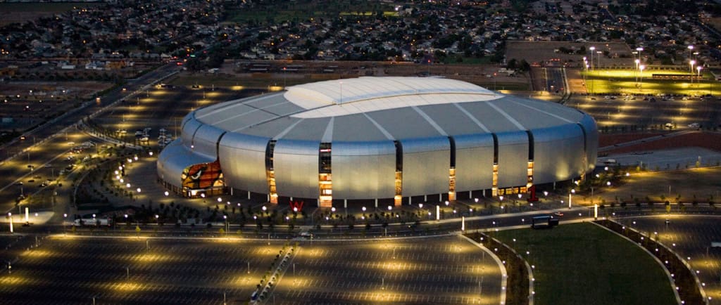 Aerial view of a large, illuminated stadium at twilight with surrounding parking areas and approaching roadways.