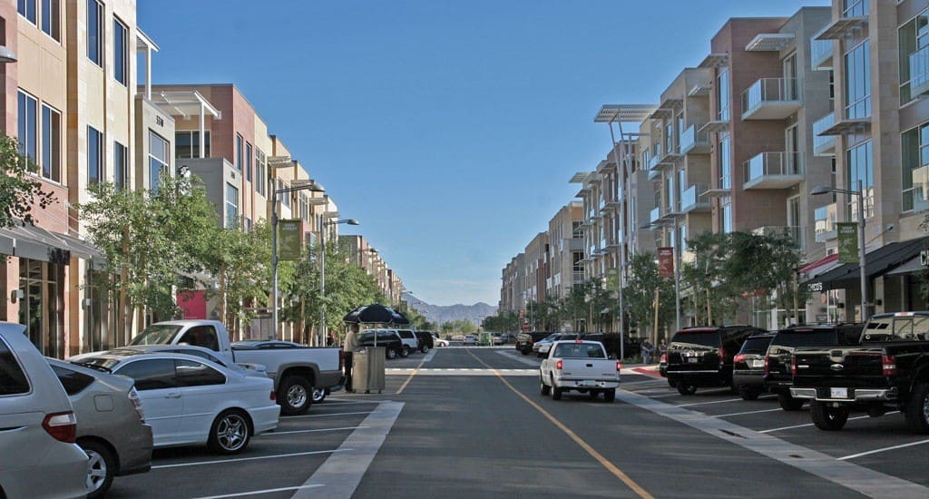 A broad street flanked by multi-story residential buildings with retail spaces on the ground floor, cars parked on both sides, and mountains visible in the distance under a clear sky.