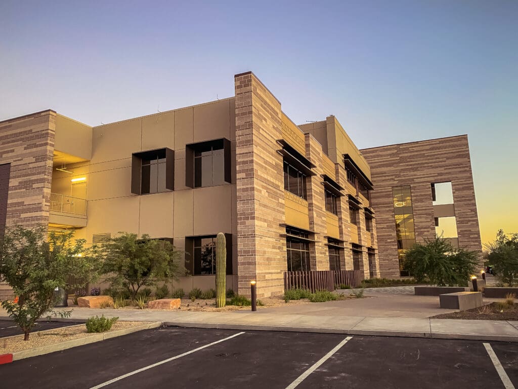 A modern office building with a stone and stucco facade illuminated by the warm glow of a setting sun, featuring ample windows and a landscaped front area, with an empty parking lot in the foreground.