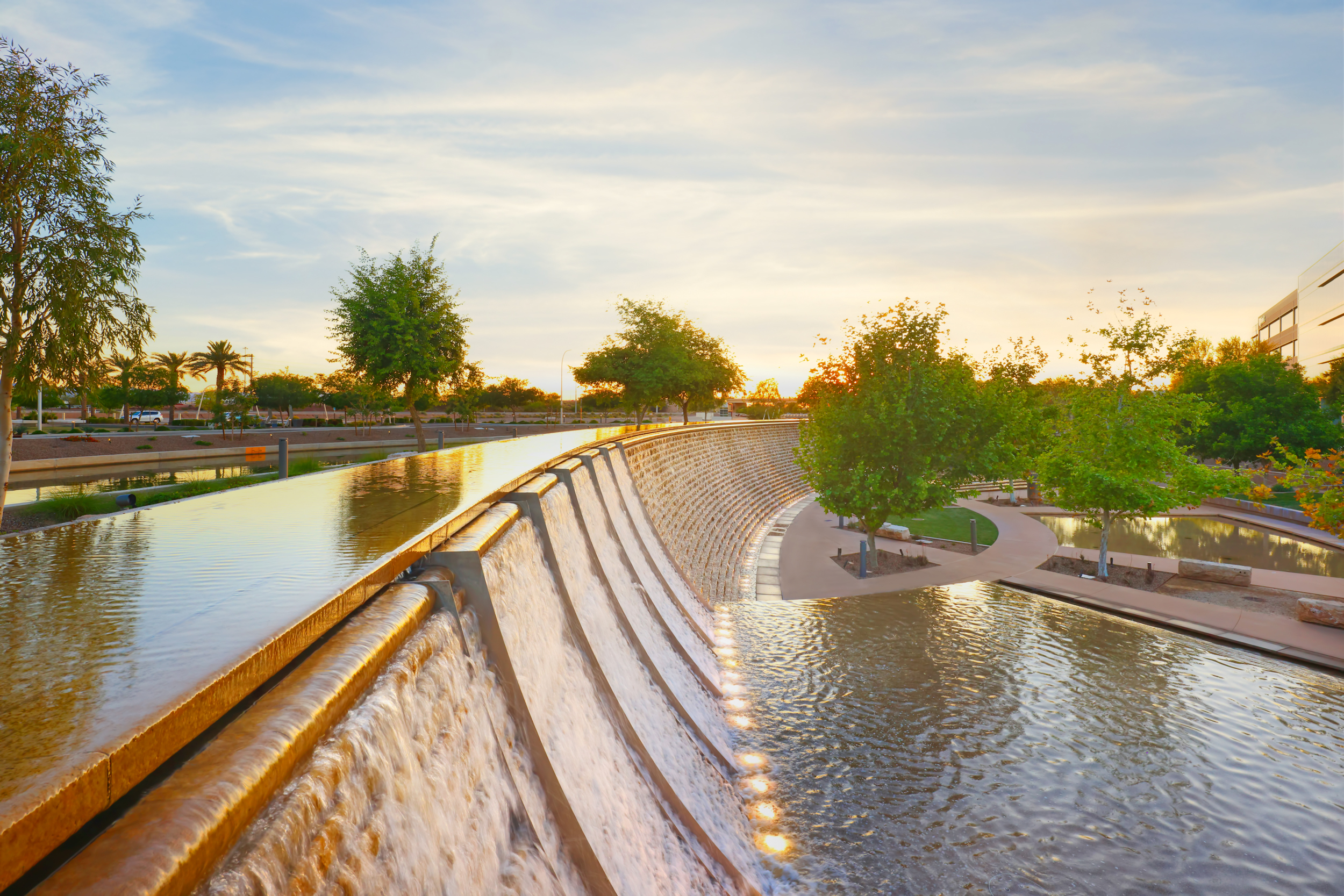 Sunset over a tranquil urban canal with flowing water, lighted paths, and lush greenery on both sides.