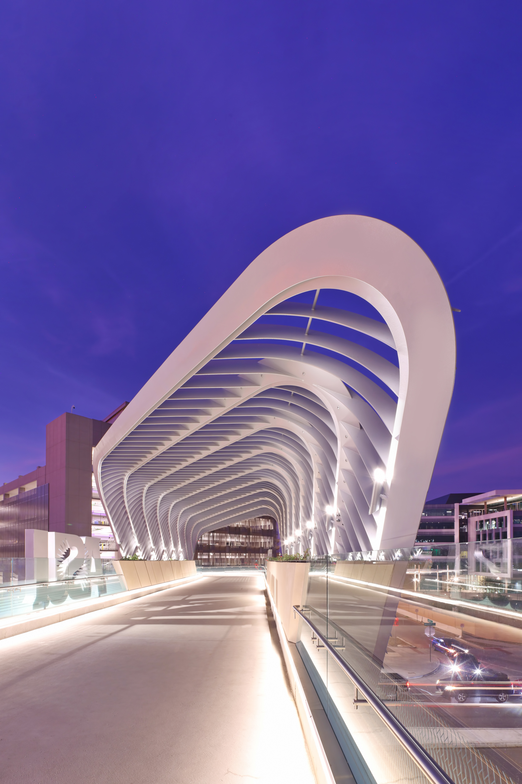 Modern bridge with a sleek, white, arching design and illuminated walkway at twilight, set against a purple sky.