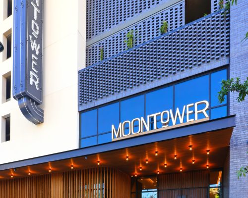 Modern building entrance with a sign reading "moontower," featuring a vertical neon sign, wood paneling, and string lights under an overhang.