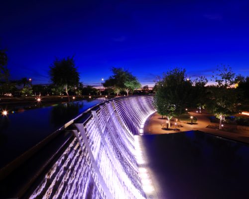 A nighttime view of an illuminated waterfall along a pathway, flanked by trees under a deep blue sky.