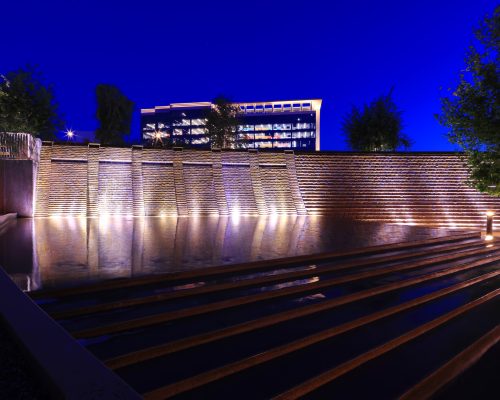 Illuminated curved water feature with lights at night in front of a modern building with a starry sky above.