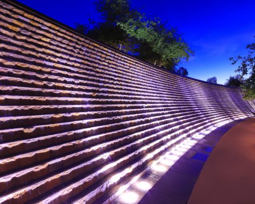 Curved stone wall illuminated by purple and white lights at night, creating long shadows with trees in the background.