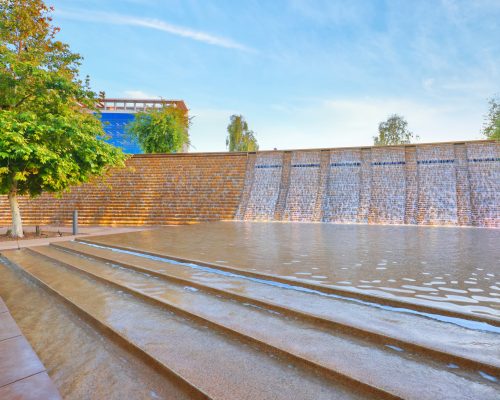 A serene water feature with a cascading waterfall wall in an urban park, surrounded by trees, under a clear blue sky.