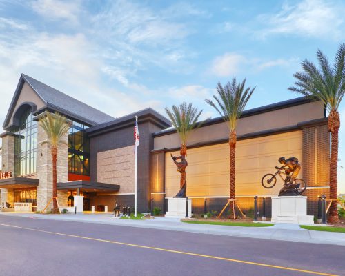 Modern commercial building with large glass windows and a motorcycle statue at the entrance, flanked by tall palm trees under a clear sky.