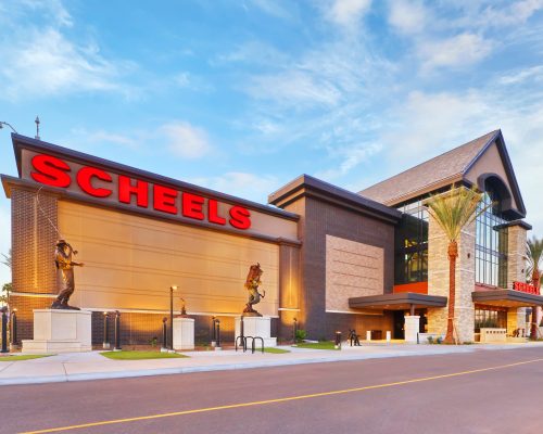 Exterior of a scheels store at dusk, featuring large red signage, statues on facade, and a clear sky.