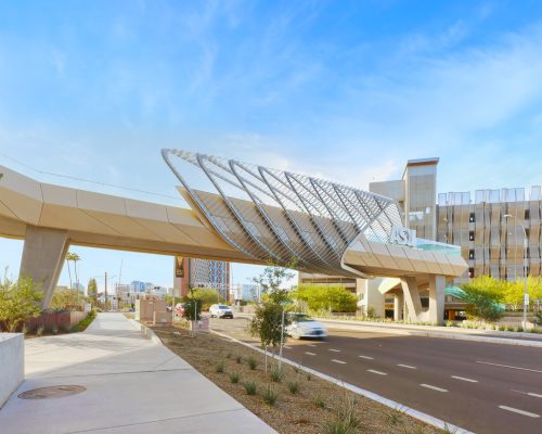 Modern pedestrian bridge with a distinctive spiral mesh design over a city street, flanked by urban buildings under a clear blue sky.