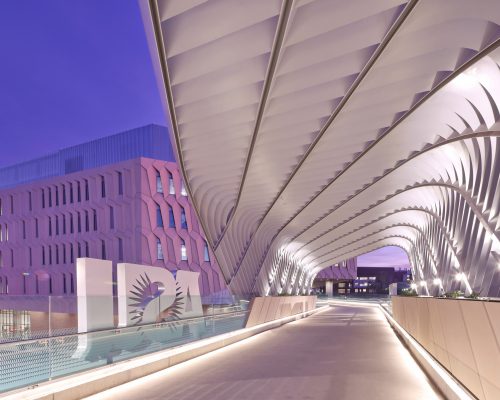 Modern architectural bridge with undulating white canopy and illuminated walkway at twilight, with pink-tinged buildings in the background.