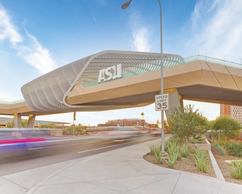 Pedestrian bridge with asu logo extending over a busy roadway with blurred vehicle lights at dusk, located in a suburban area with clear skies.