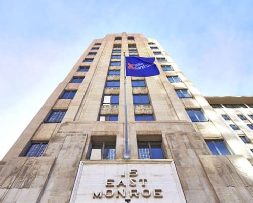 A tall beige building with a "bank of america" sign and "5 east monroe" inscription on the facade, viewed from a low angle against a clear blue sky.
