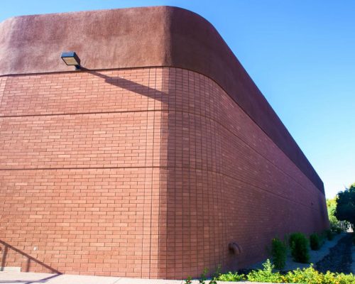 A brown brick building with a curved wall under a clear blue sky, casting a shadow from a mounted outdoor light.