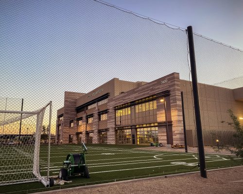 A soccer goal and a maintenance cart on an artificial turf field with a net overhead, in front of a modern building at dusk.