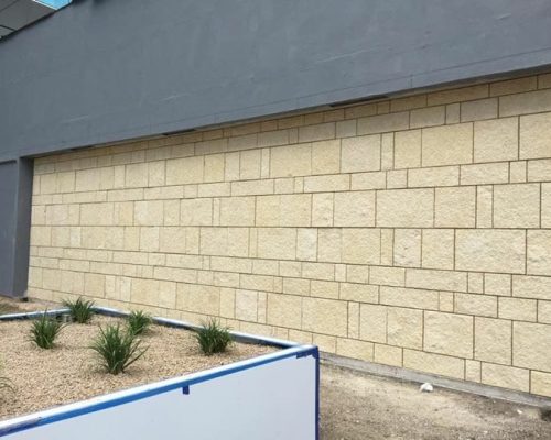 A beige brick wall beside a grey garage door with small plants at the base and a blue barrier in front.