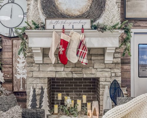 A cozy living room decorated for christmas with stockings hung on the mantle, a festive wreath above the fireplace, and candles inside the hearth.