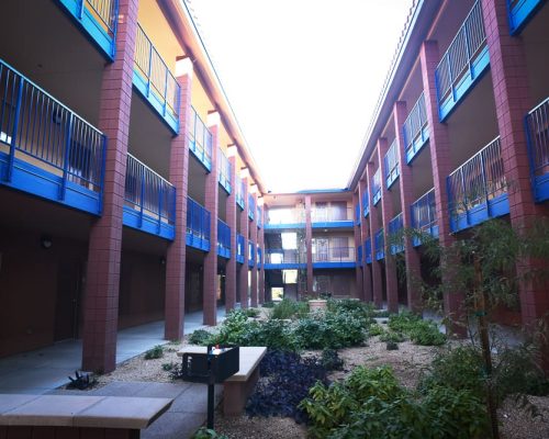 An empty courtyard within a residential building featuring two stories with blue railings and red exterior walls, surrounded by small green shrubs and trees under a bright sky.