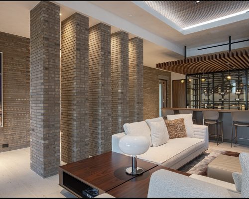 Residential modern living room with exposed brick columns, a sectional sofa, and a view into a kitchen with bar stools.