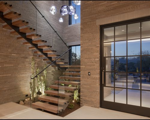 Residential modern staircase in a well-lit home entryway with glass pendant lights and a brick wall.