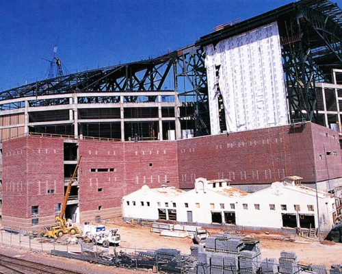Construction site of a large stadium with steel framework partially completed and brickwork structures visible at the foreground.