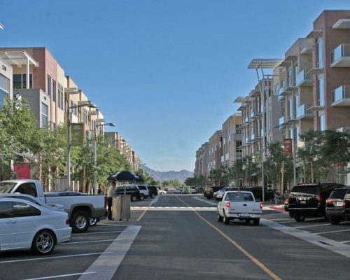 A broad street flanked by multi-story residential buildings with retail spaces on the ground floor, cars parked on both sides, and mountains visible in the distance under a clear sky.