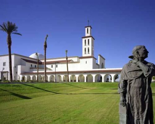 A statue of a person stands in the foreground, gazing toward a white building with a bell tower and multiple arches, surrounded by palm trees under a clear blue sky.